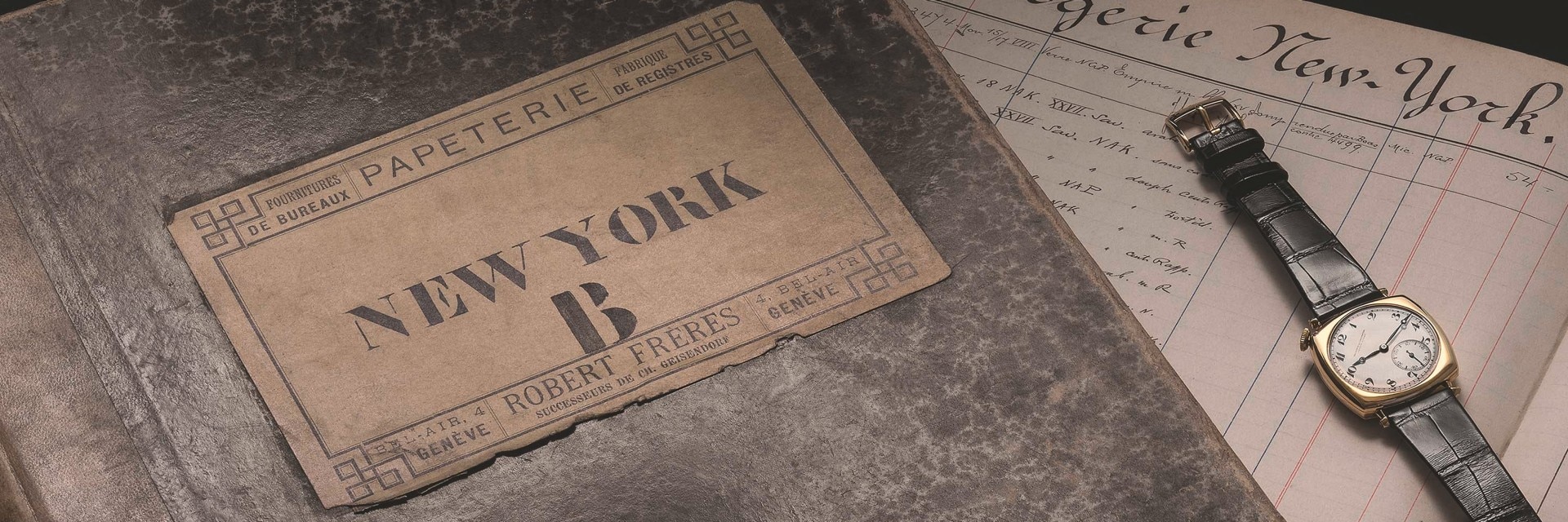 A vintage book labeled 'NEW YORK B', next to an old-fashioned wristwatch with a leather strap, placed on top of an old document with handwritten text