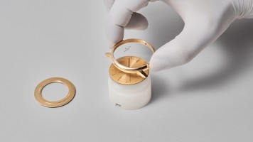 Watchmaker's gloved hand assembling a gold watch case on a white surface.