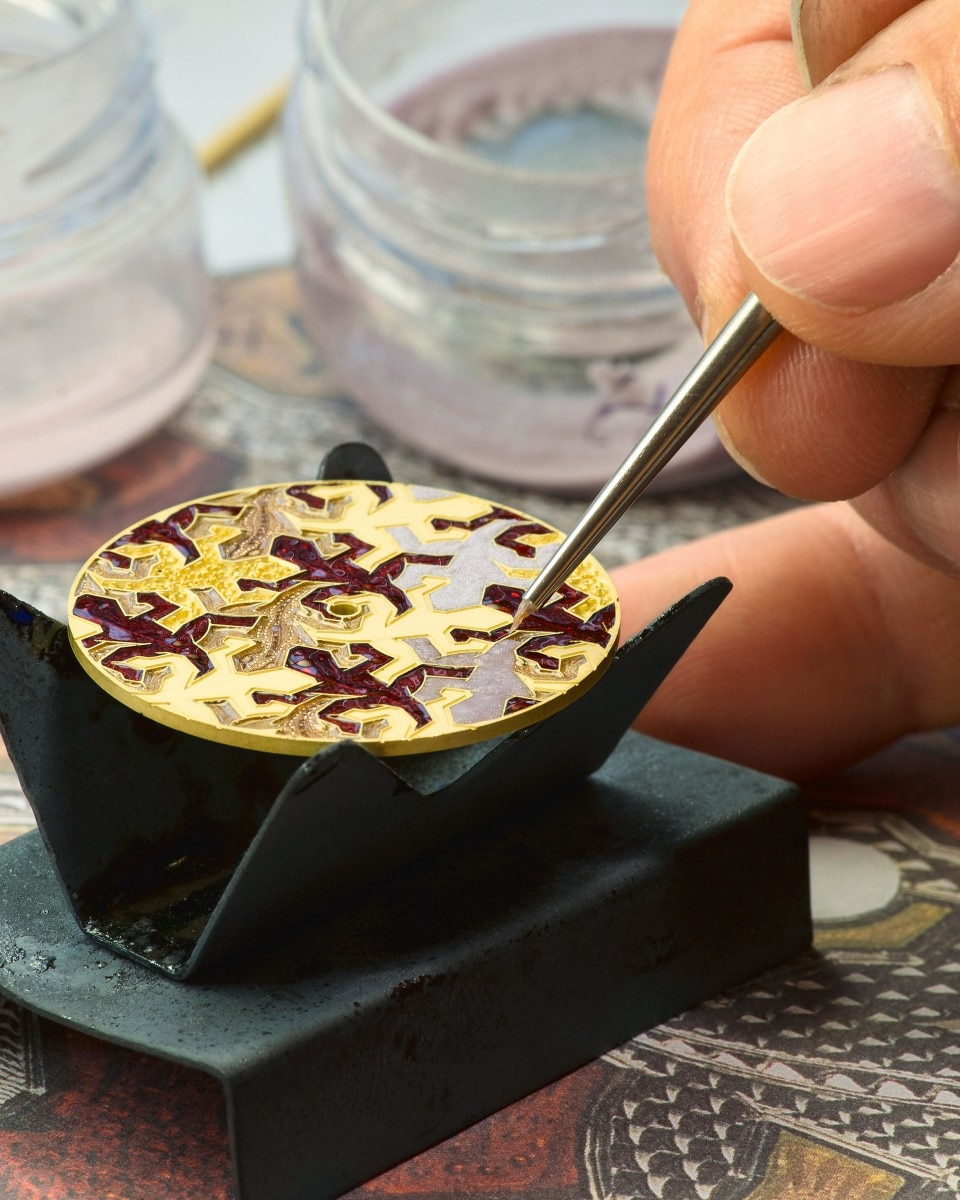 Artisan using a fine brush to apply enamel colors onto a patterned watch dial.
