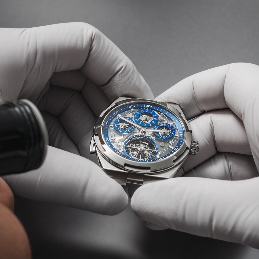 Watchmaker inspecting a blue-dial luxury watch with gloved hands and a loupe.