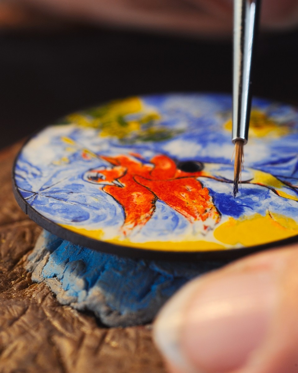 Close-up of a hand applying bright colors to a watch dial with a floral and bird motif.