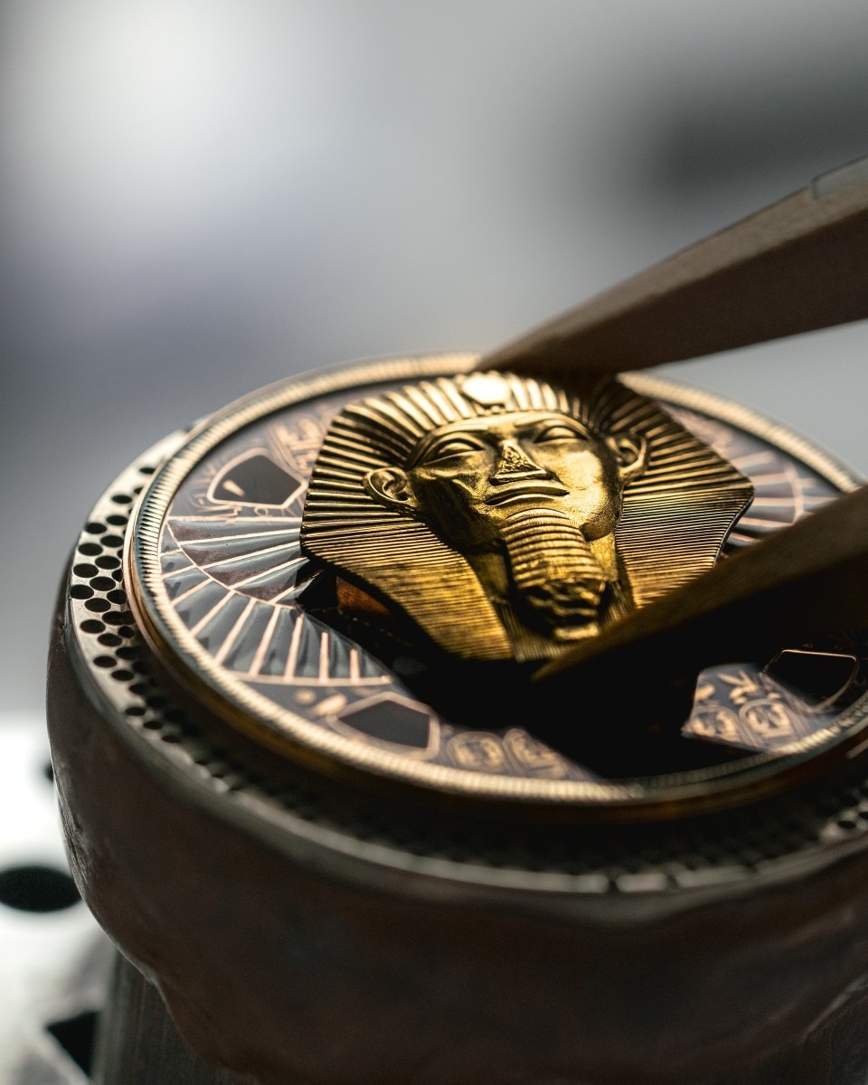 Watch dial being assembled with a golden sculpted figure using tweezers.