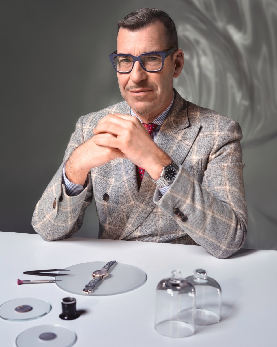 A man in a light suit sits at a table with a watch and tools in front of him.