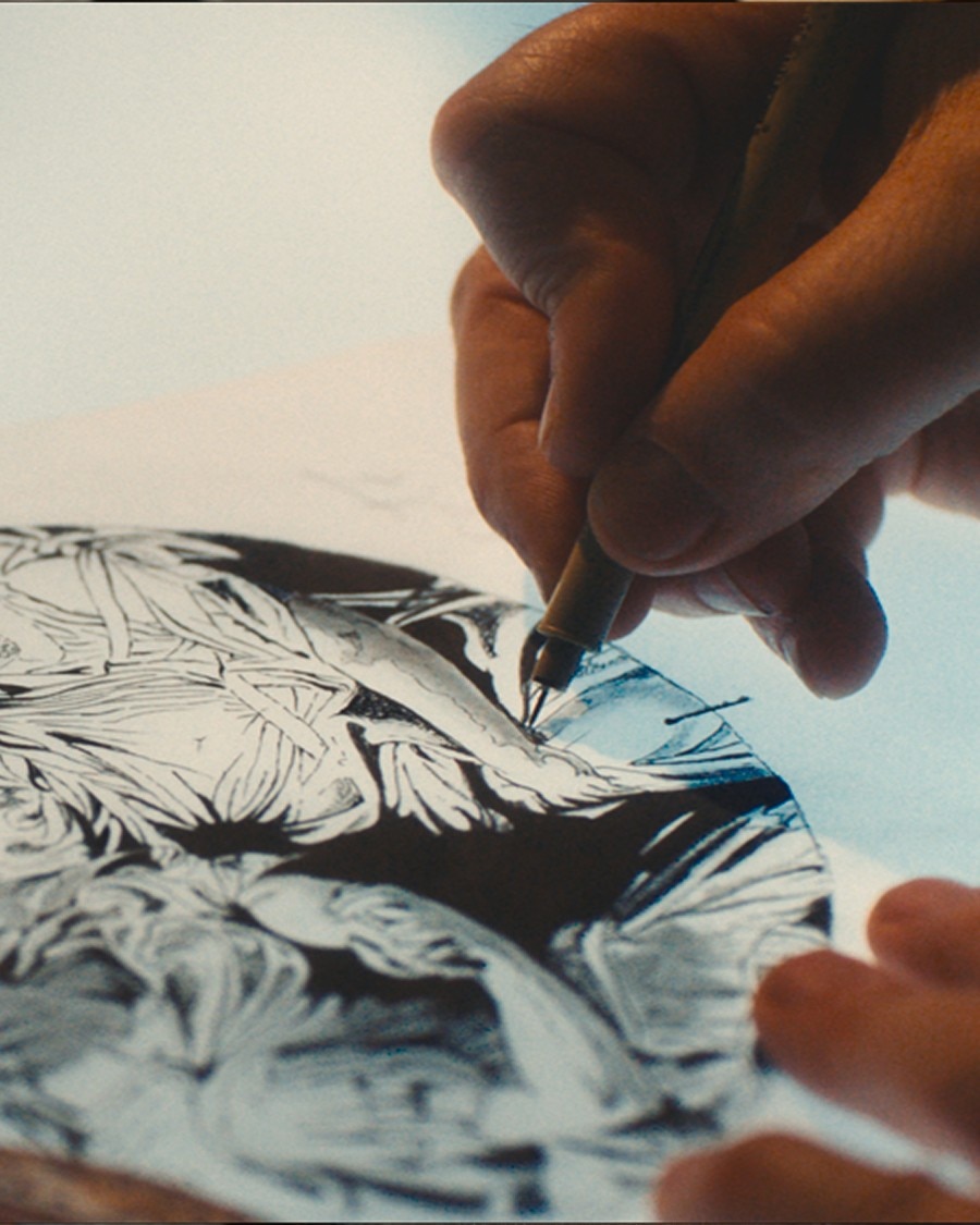Artisan drawing detailed watch dial design with a pen on tracing paper.