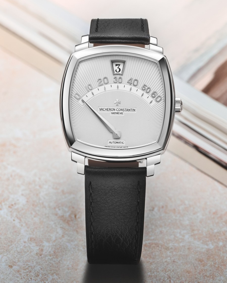 Wristwatch with a black leather strap, a silver watch face featuring an arched scale from 0 to 60 for minutes, and a small window at the top displaying the hour in numerical format.