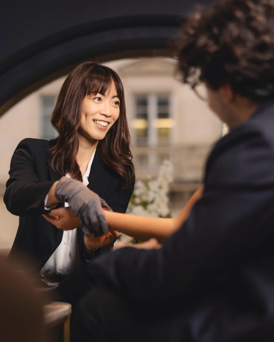 Une femme en tailleur noir avec des gants examine une montre de luxe en compagnie d’un homme, dans un cadre professionnel.