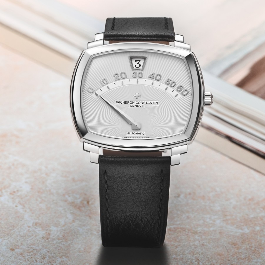 Wristwatch with a black leather strap, a silver watch face featuring an arched scale from 0 to 60 for minutes, and a small window at the top displaying the hour in numerical format.
