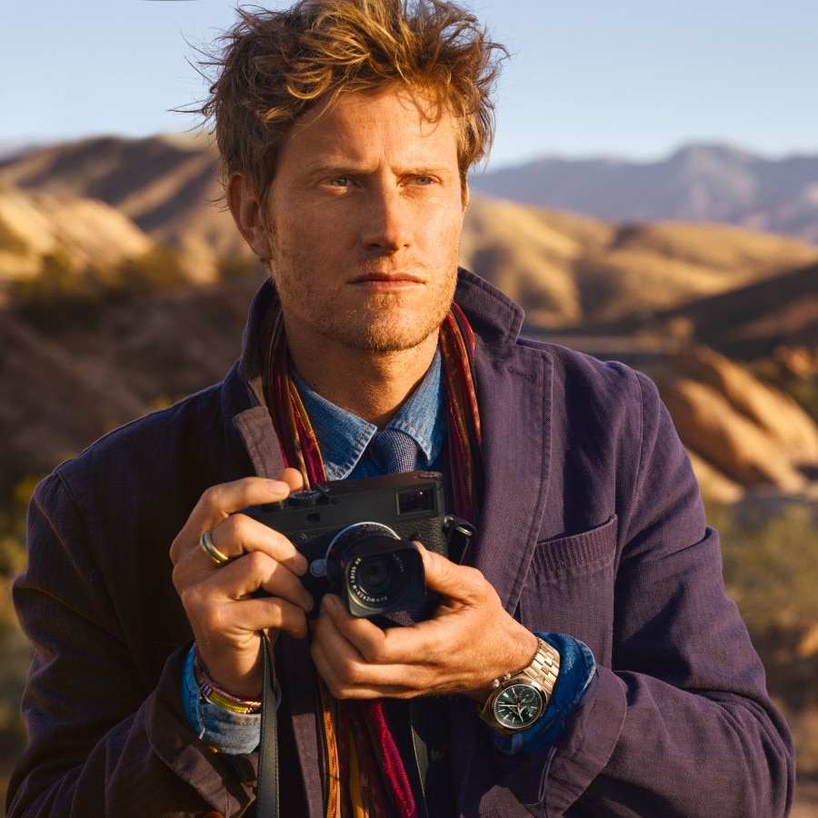 Man holding a camera in a desert landscape, wearing a purple blazer and a luxury wristwatch.