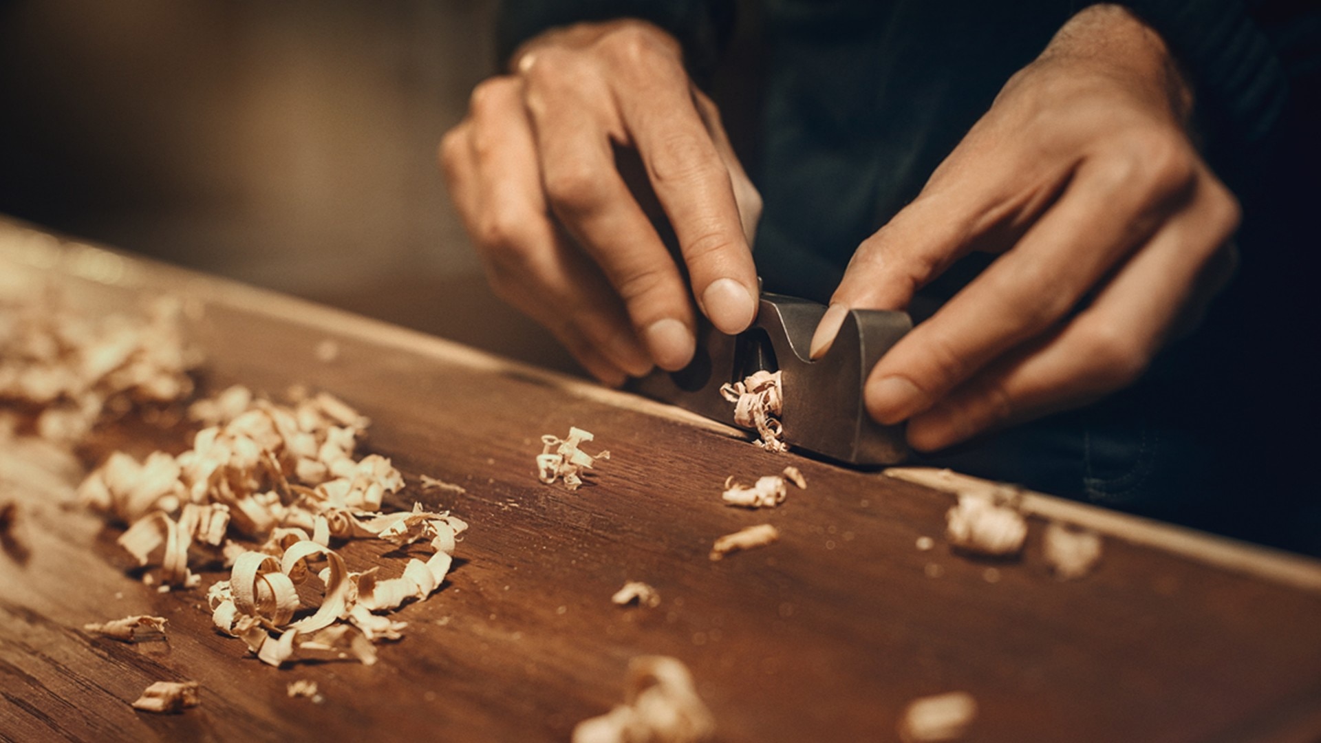 Close-up of hands carving wood shavings with a chisel on a workbench.