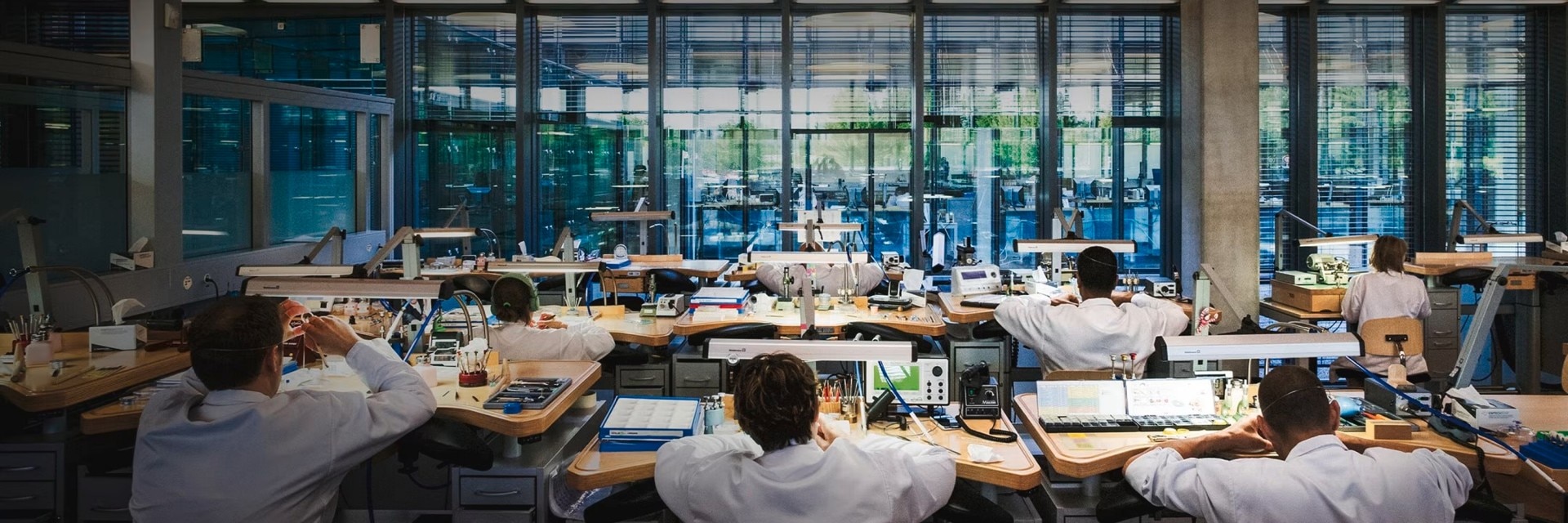 Wide view of a watchmaking workshop with artisans working at desks in a modern, glass-walled space.