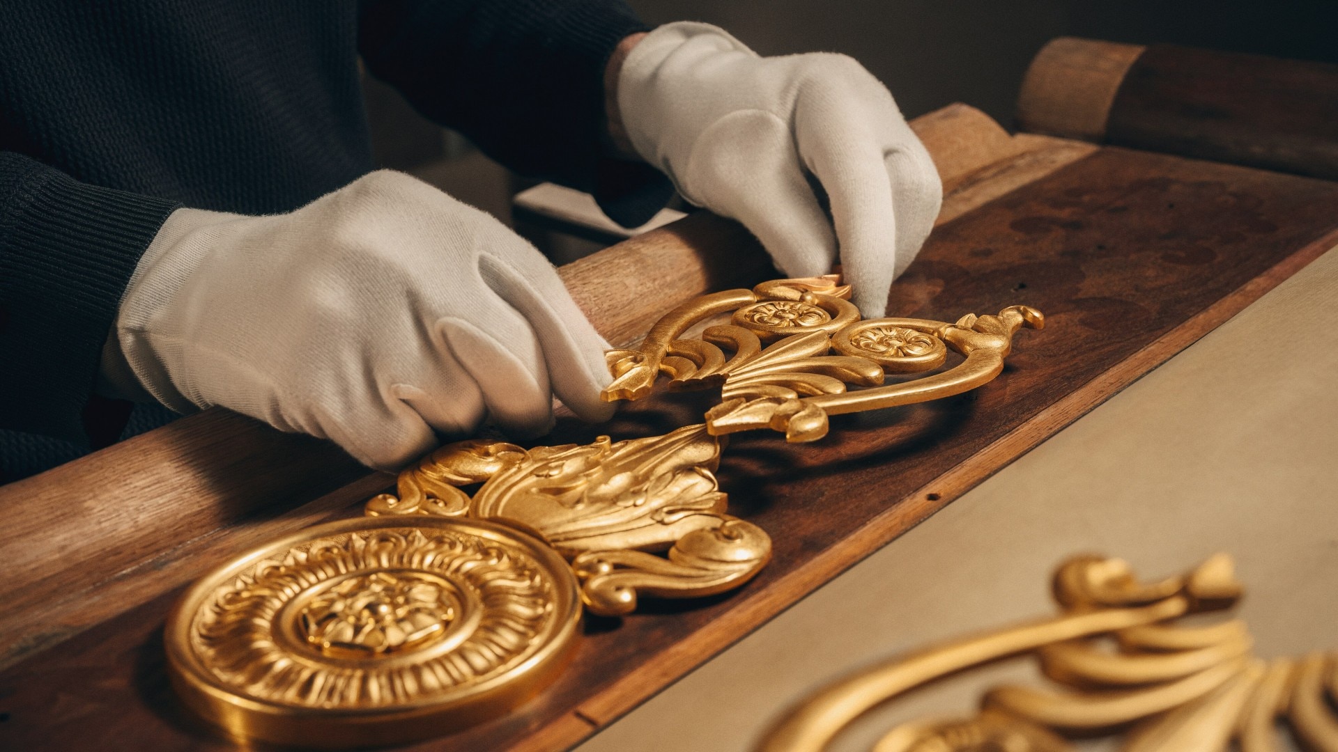 Close-up of a gloved hands restoring or assembling intricate golden clock components on a wooden surface.