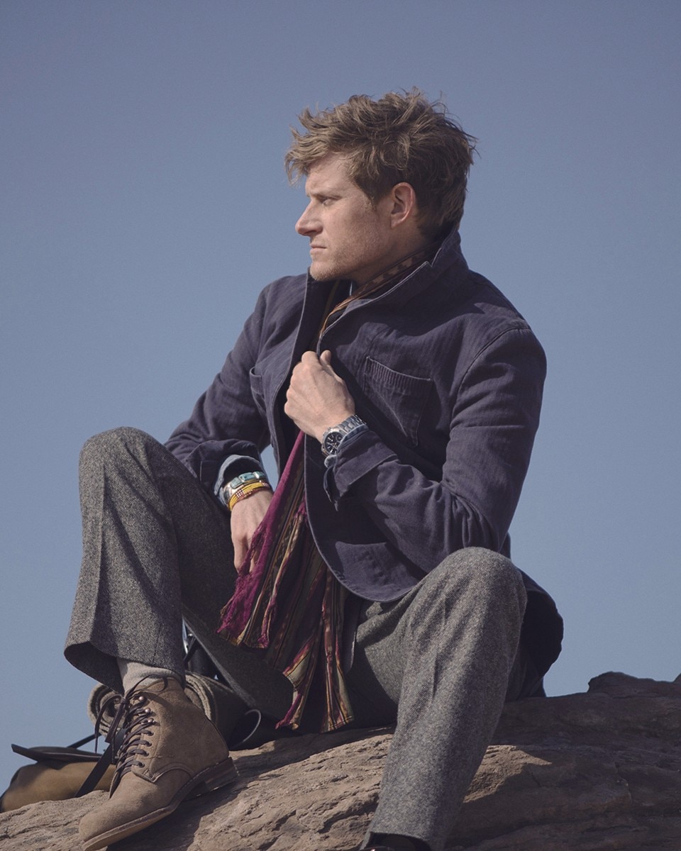 Uomo in abbigliamento outdoor seduto su una roccia, che guarda di lato mentre tiene in mano un orologio.