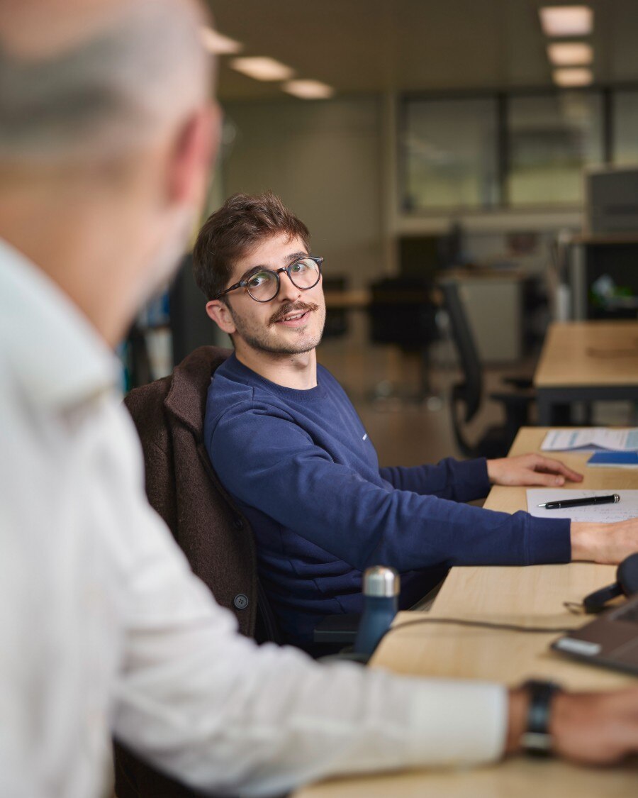 Two men having a conversation in an office, one seated at a desk and smiling.