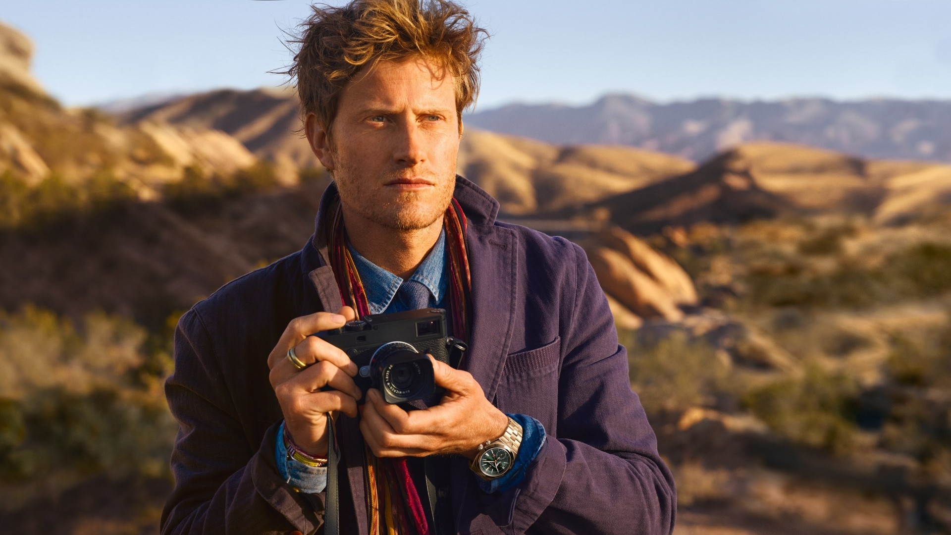 Man holding a camera in a desert landscape with mountains in the background.