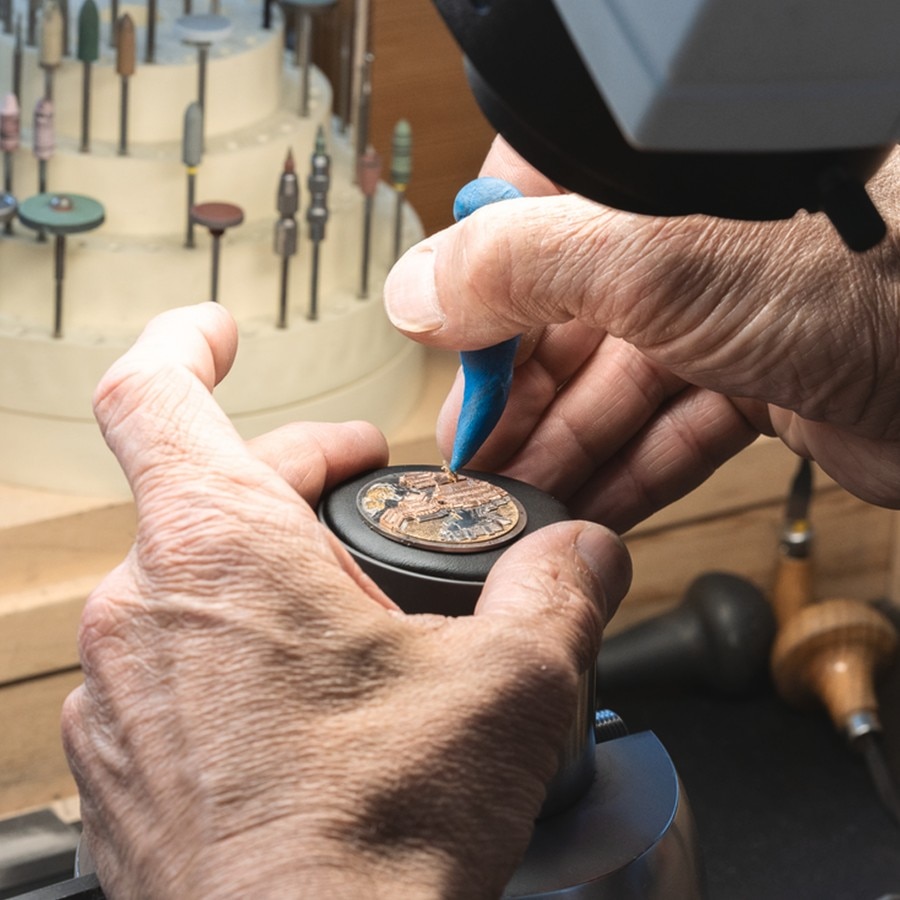 Watchmaker working on a detailed watch dial using precision tools under a microscope, with various tools in the background.