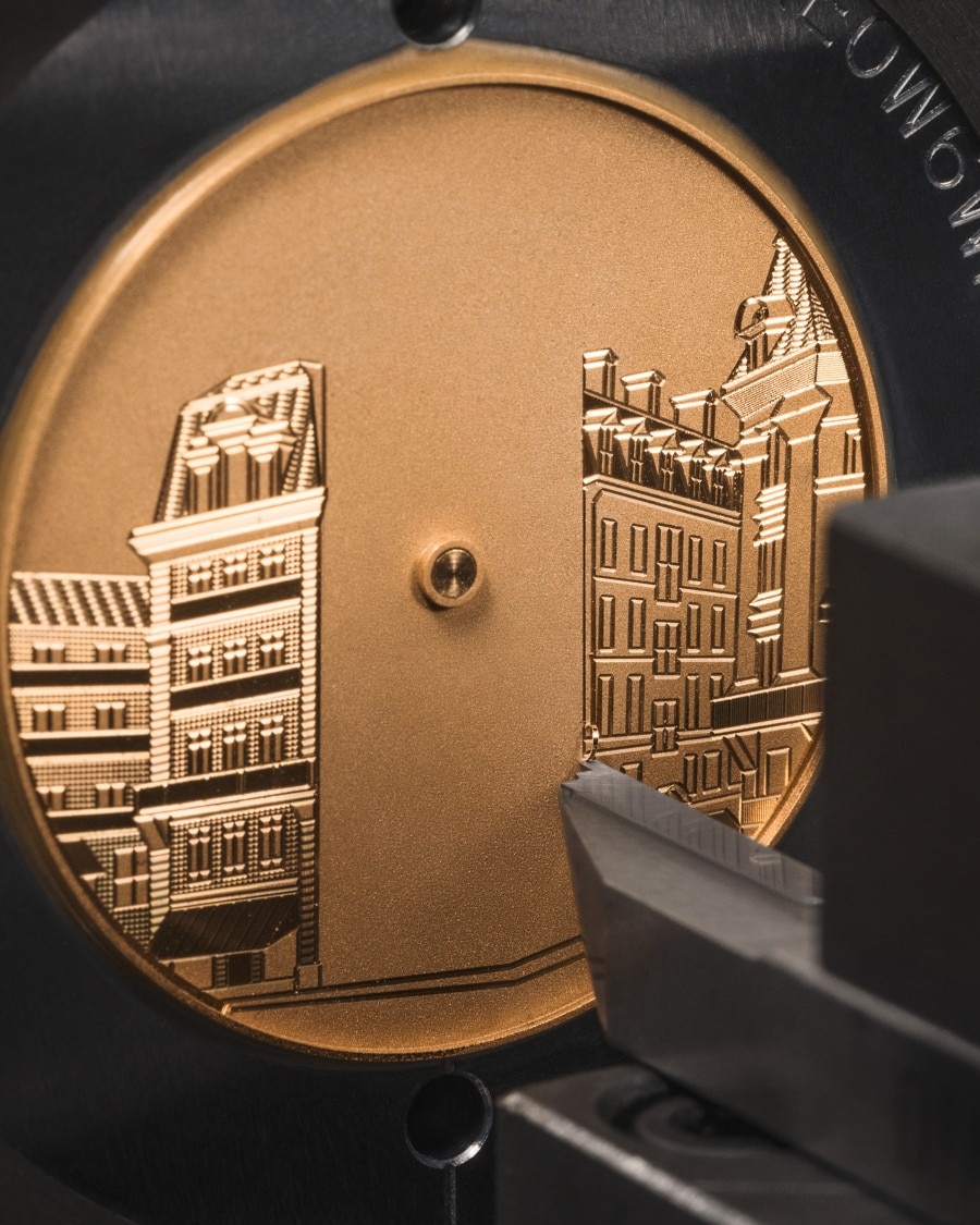 Engraved cityscape in progress on a bronze-colored watch dial, held in a precision machine.