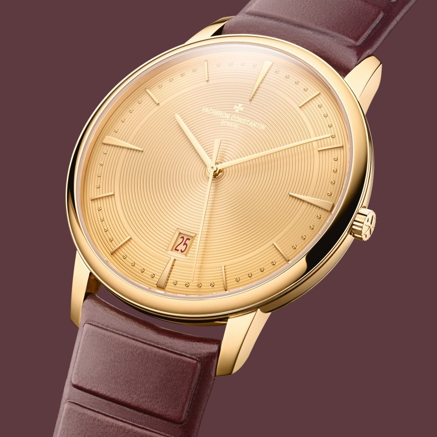 Close-up of a gold Vacheron Constantin watch with a brown leather strap, set against a burgundy background.