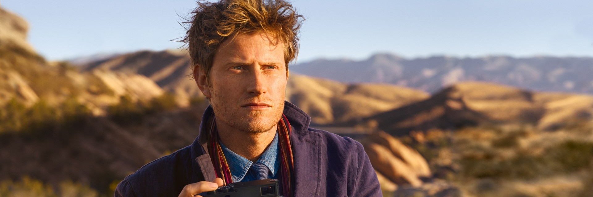 Homme tenant un appareil photo dans un paysage désertique avec des montagnes en arrière-plan.