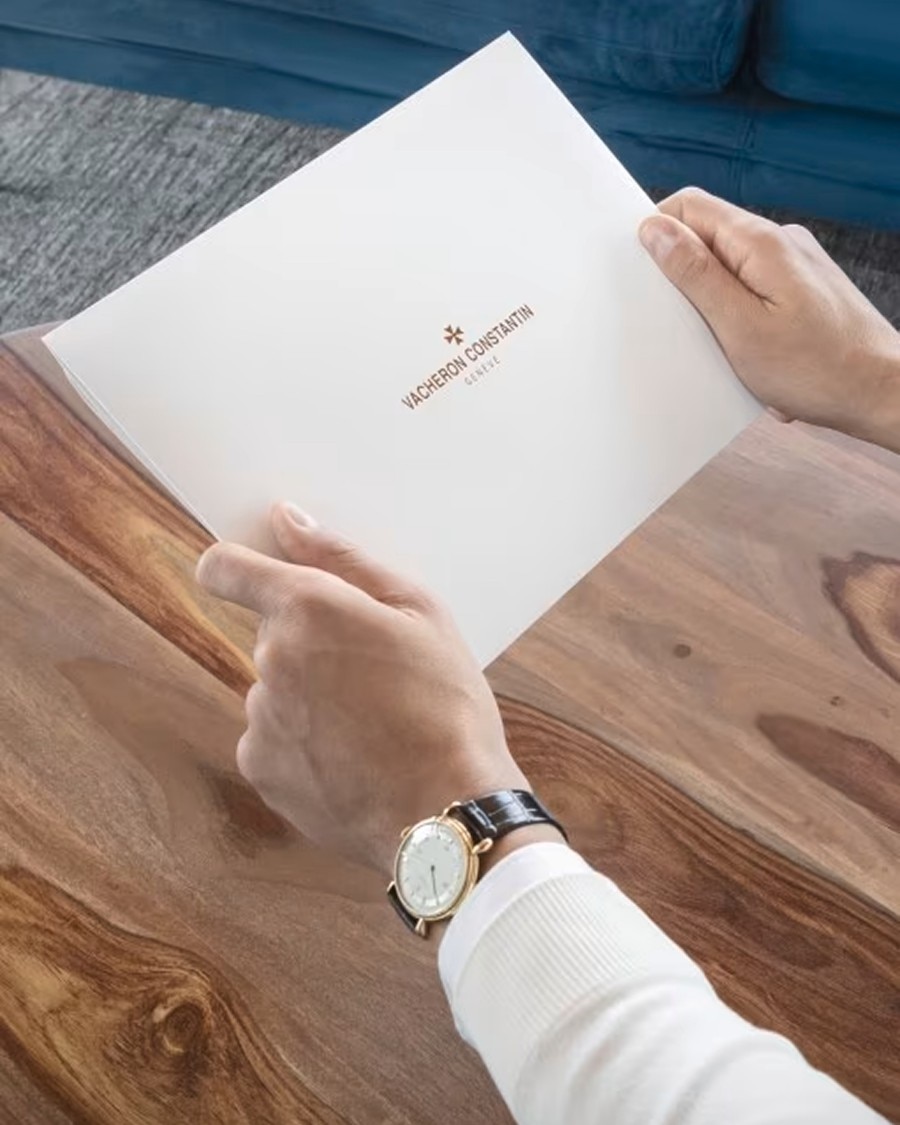 Man wearing a wristwatch while holding a white envelope at a wooden table.