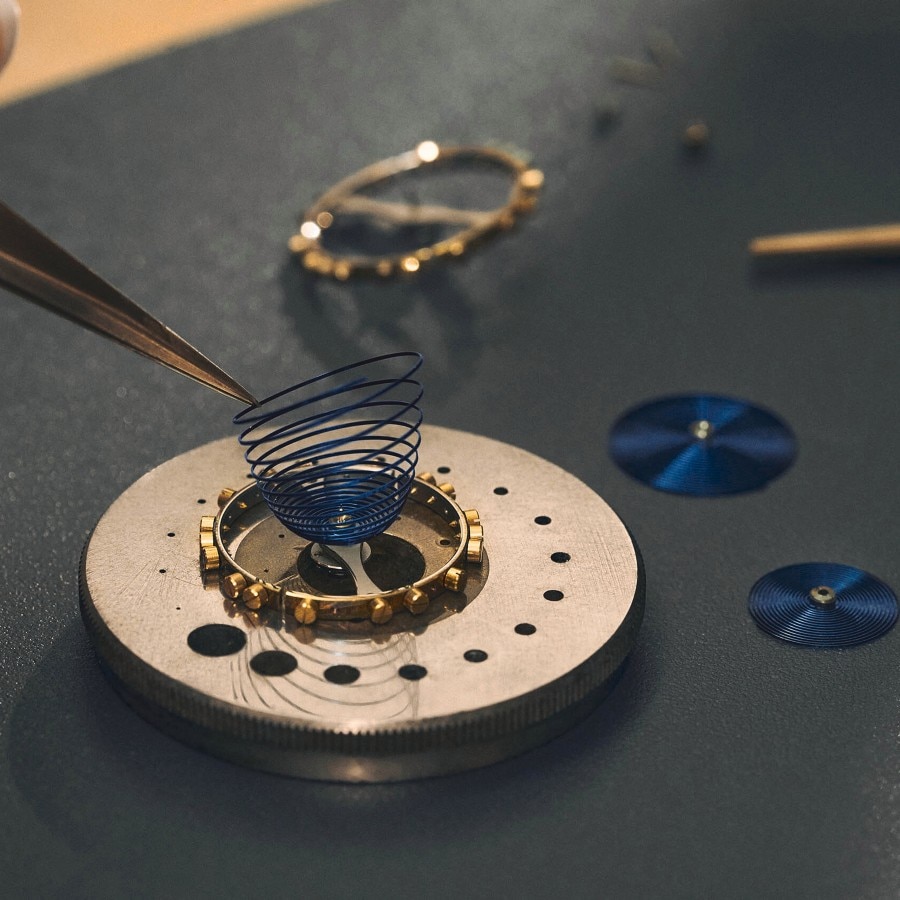 Watchmaking components on a workbench, with tweezers holding a delicate hairspring above a gear assembly.