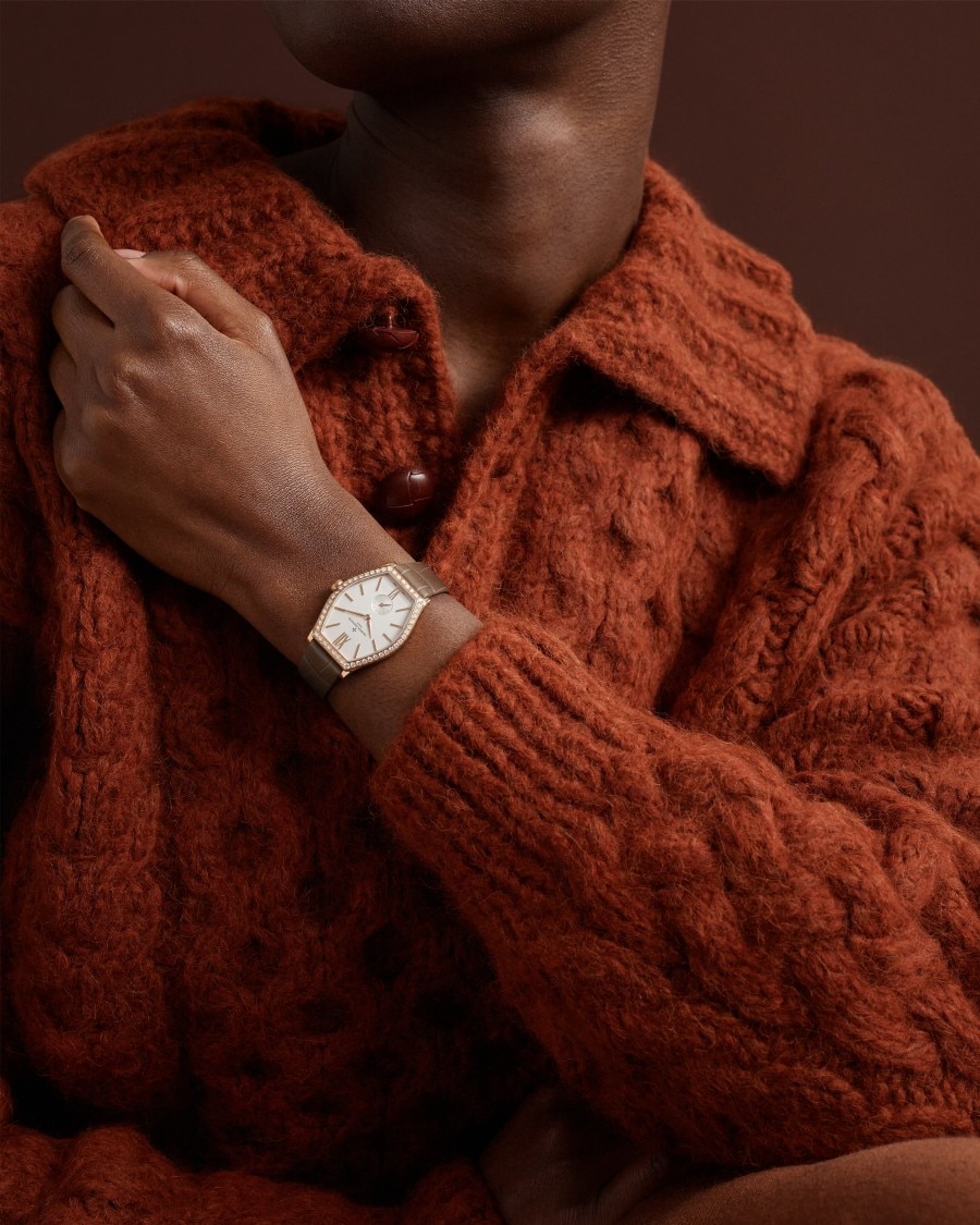 Malte Malte watch with gold case and brown leather strap on wrist in textured orange sweater.